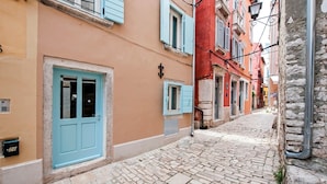 A narrow cobblestone street lined with pastel-colored buildings, featuring blue doors and shutters, and a stone wall on one side. - CASA ROCCIA BIANCA - CIELO - Two Bedroom Apartment (Rovinj)