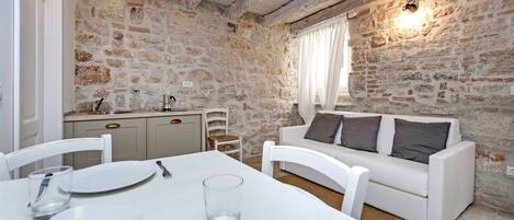 A dining area with a white table set for two, a stone wall, a white sofa with cushions, and a kitchen area with a sink and cabinets.