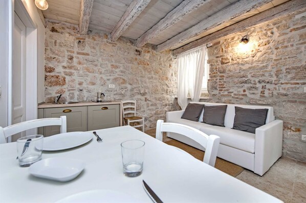 A dining area with a white table set for two, a stone wall, a white sofa with cushions, and a kitchen area with a sink and cabinets. - CASA ROCCIA BIANCA - MARE - One Bedroom Apartment (Rovinj)