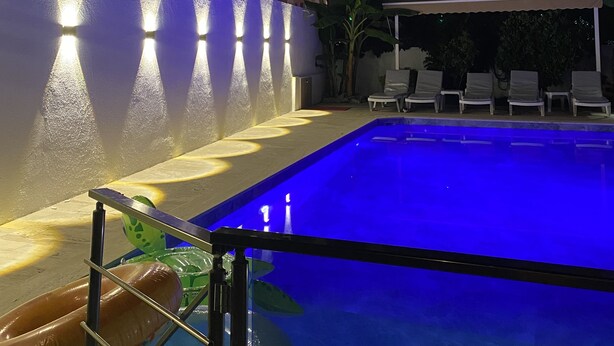 A swimming pool with a blue tint, surrounded by lounge chairs and a wall with illuminated panels. - Iberia Villas (Ortaca)