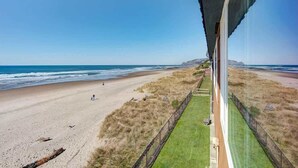 A beach view from a window overlooking a sandy beach with a grassy area and a fence. - Sea Gypsy Rentals -  Oceanfront Suite - Pool! (Lincoln City)