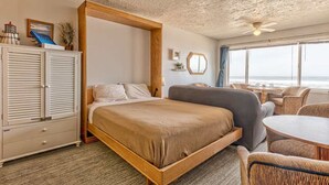 A bedroom with a large bed, a wooden wardrobe, a small table, and a view of the ocean. - Sea Gypsy Rentals -  Oceanfront Suite - Pool! (Lincoln City)