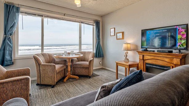 A room with a large window offering a view of the ocean, a television, and a seating area with a table and chairs. - Sea Gypsy Rentals -  Oceanfront Suite - Pool! (Lincoln City)