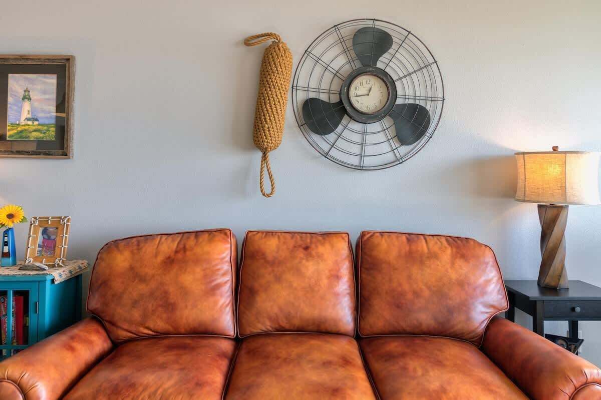 Una habitación con un sofá de cuero marrón, un ventilador de pared, un cuadro enmarcado y una lámpara sobre una mesita.