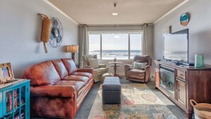 A living room with a leather sofa, armchairs, a coffee table, and a TV on a wooden cabinet. A large window offers a view of the ocean. - Oceanfront Suite - Top Floor - Pool and Sauna - Sleeps 4! (Lincoln City)
