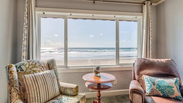 A room with a large window overlooking a beach, a round table with a vase, and two upholstered chairs. - Oceanfront Suite - Top Floor - Pool and Sauna - Sleeps 4! (Lincoln City)