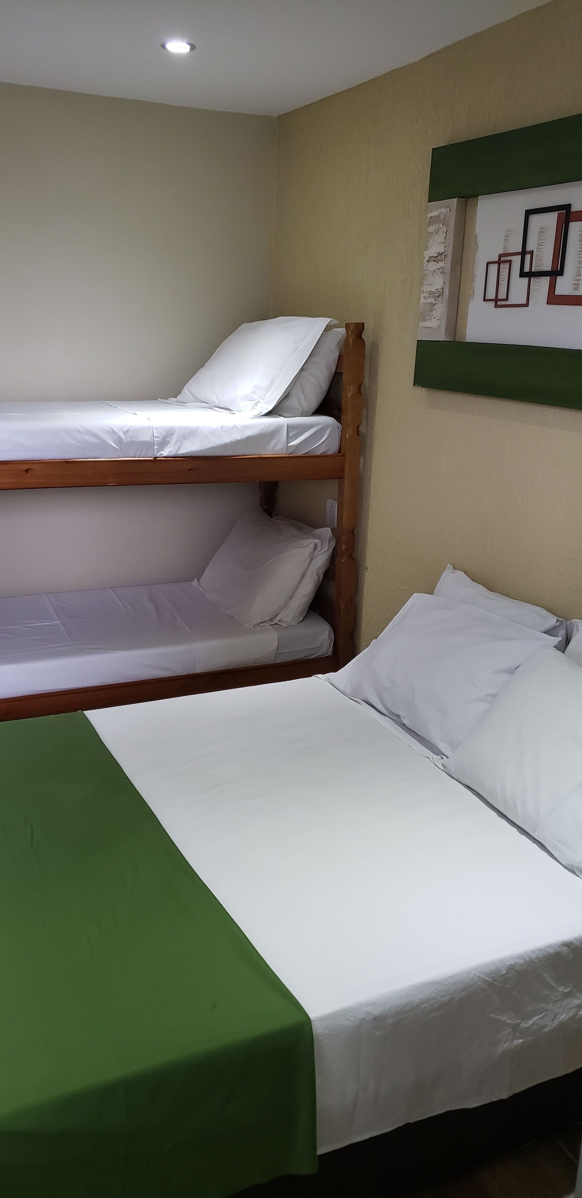 A bunk bed with white bedding and a green bedspread, a single bed with white bedding, and a wall-mounted shelf with framed pictures.