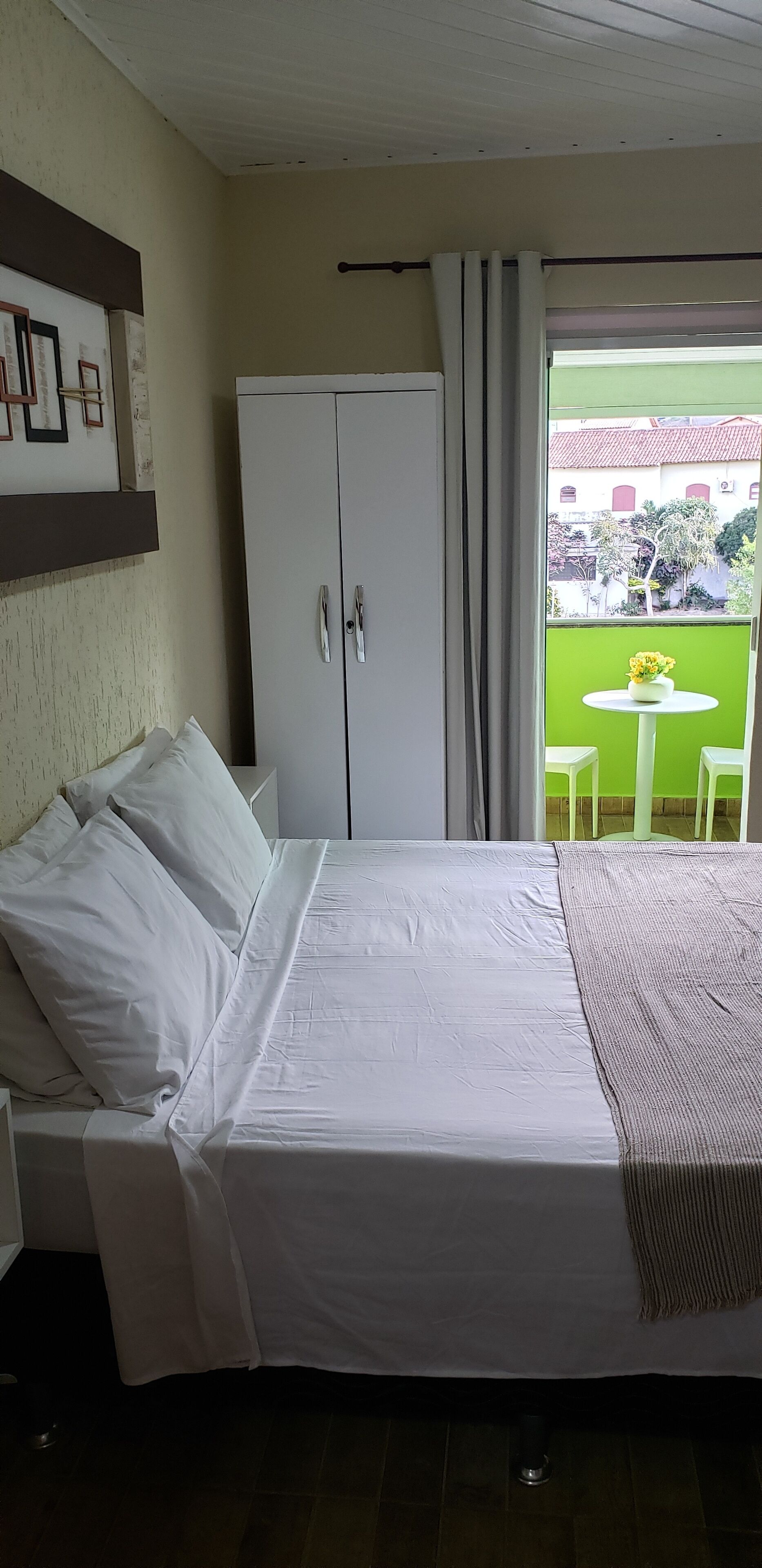 Un dormitorio con cama, armario, una mesita con un florero y vista a un área verde exterior.