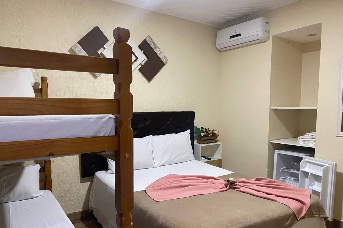 A hotel room with a bunk bed, a single bed with a pink blanket, a bedside table with a plant, and a white wardrobe.
