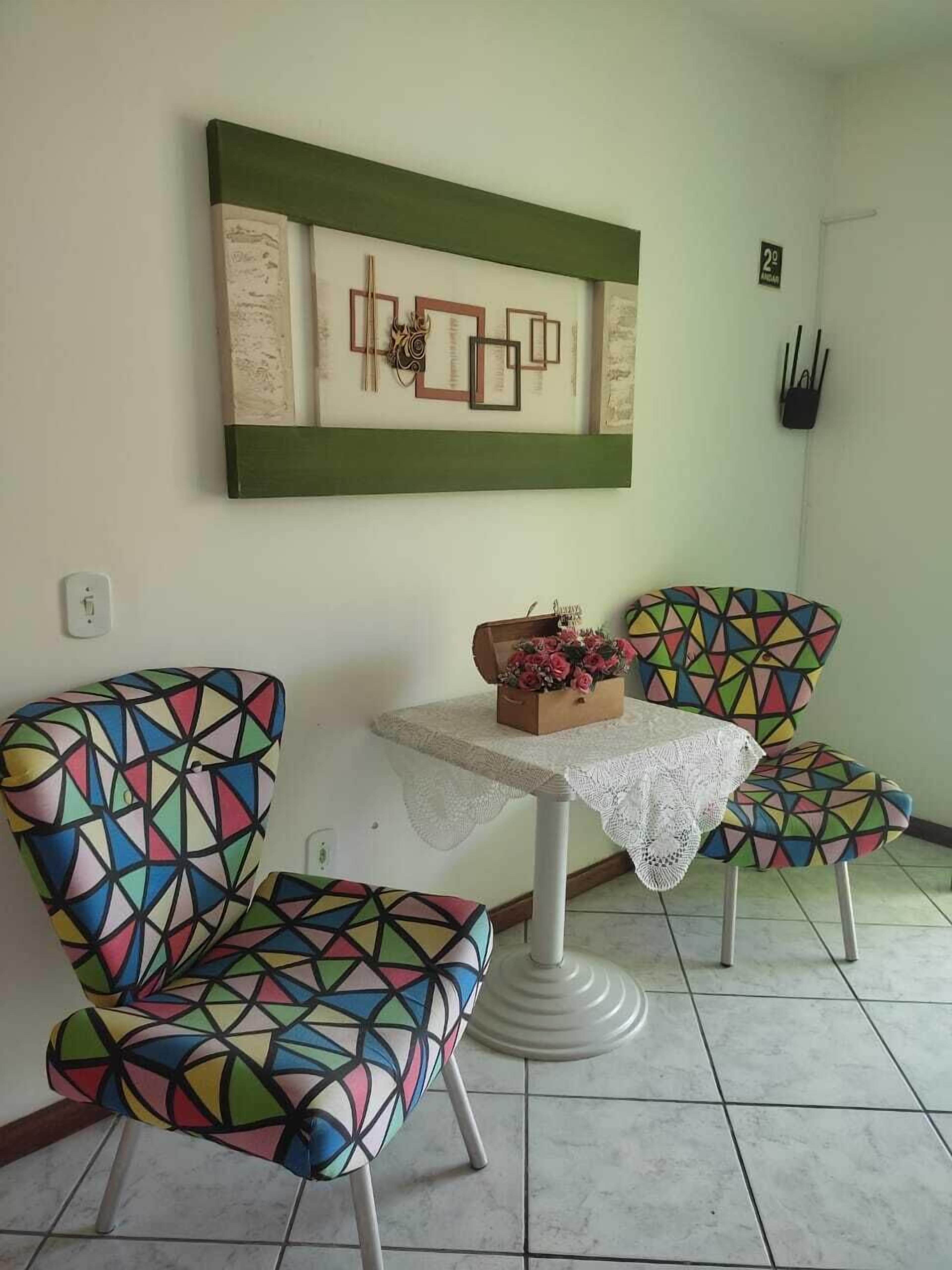 A room with two colorful chairs, a small table with a lace tablecloth, and a wall-mounted artwork.