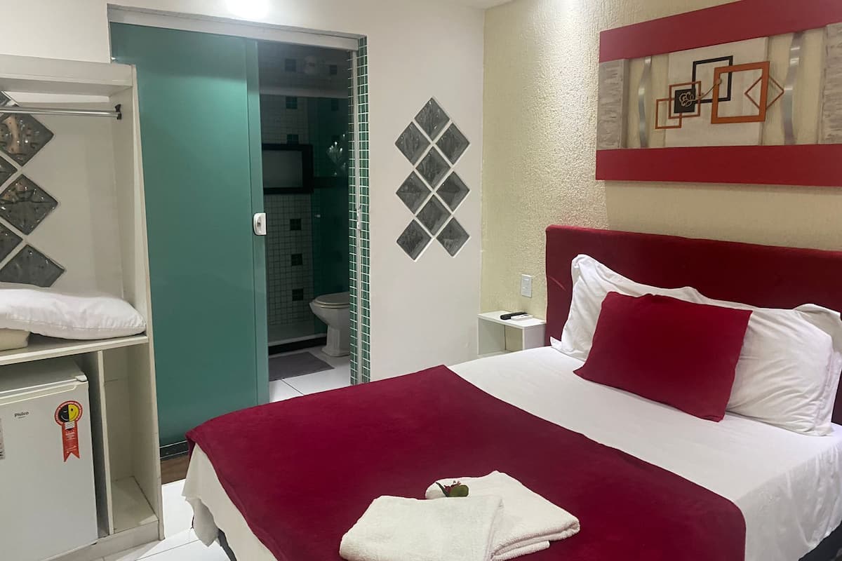 A hotel room with a bed, a nightstand, a red headboard, a white pillow, a red pillow, a white towel, a small table, and a mirror.