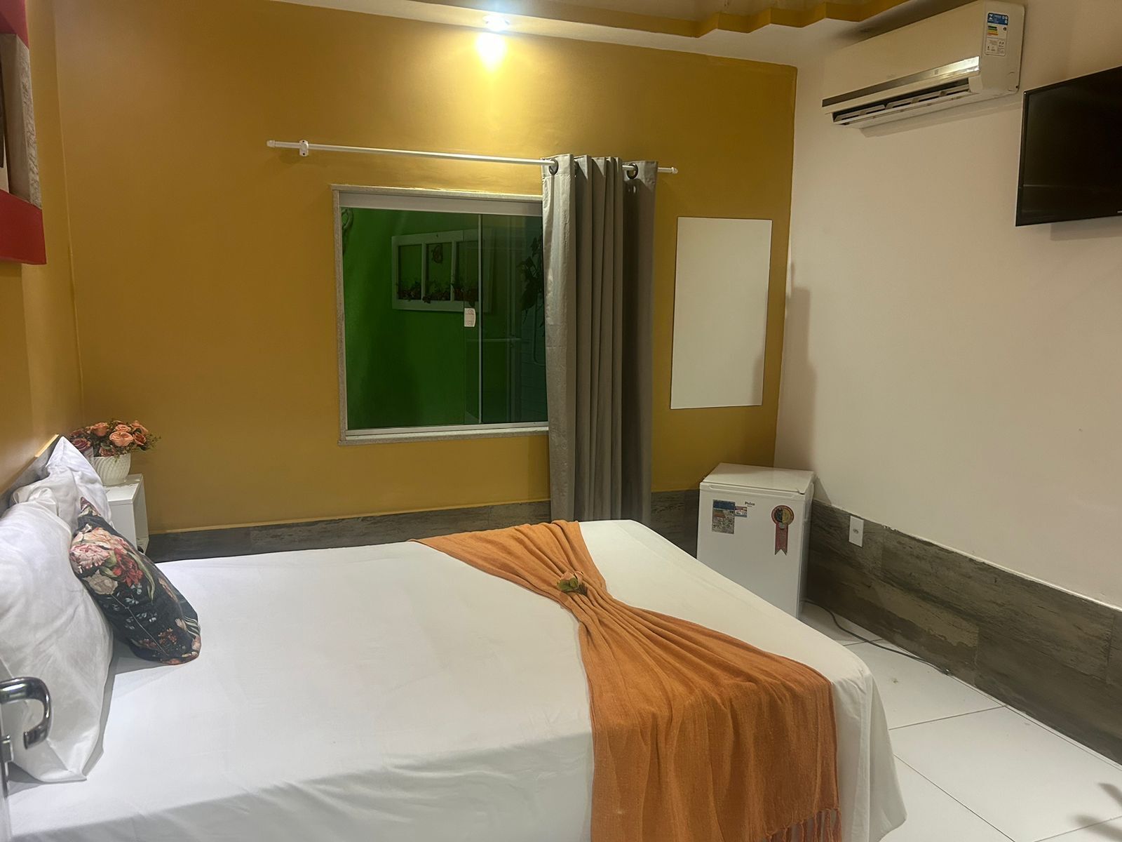 Habitación de hotel con cama, ventana con cortinas, televisor montado en la pared, aire acondicionado y una mesita con un jarrón de flores.