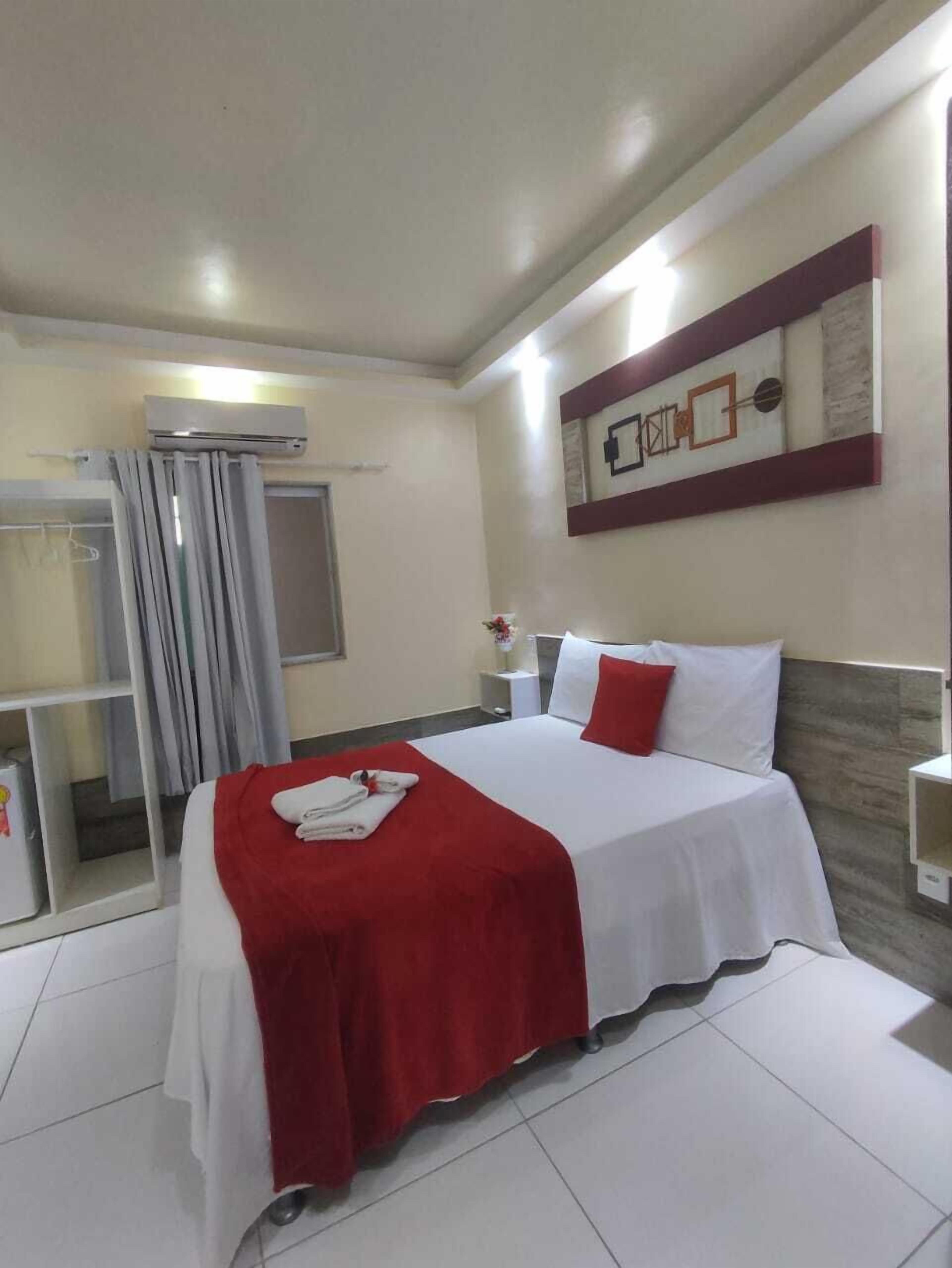 Une chambre d’hôtel avec un lit, un plaid rouge, un oreiller blanc, une table de chevet, une œuvre d’art accrochée au mur et une fenêtre avec des rideaux.