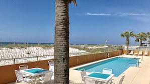 Zona junto a la piscina con sillas reclinables blancas y una piscina, con vistas a una playa con dunas de arena y un cielo despejado.