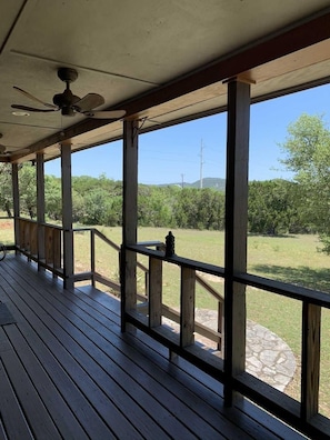 A covered outdoor deck with a ceiling fan, wooden railings, and a view of a grassy area with trees.