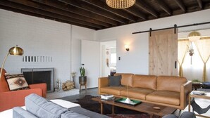 A modern living room with a leather sofa, a grey sectional, and a coffee table. There is a fireplace, a hanging light fixture, and a wooden door. - The Ridge at Joshua Tree (Joshua Tree)