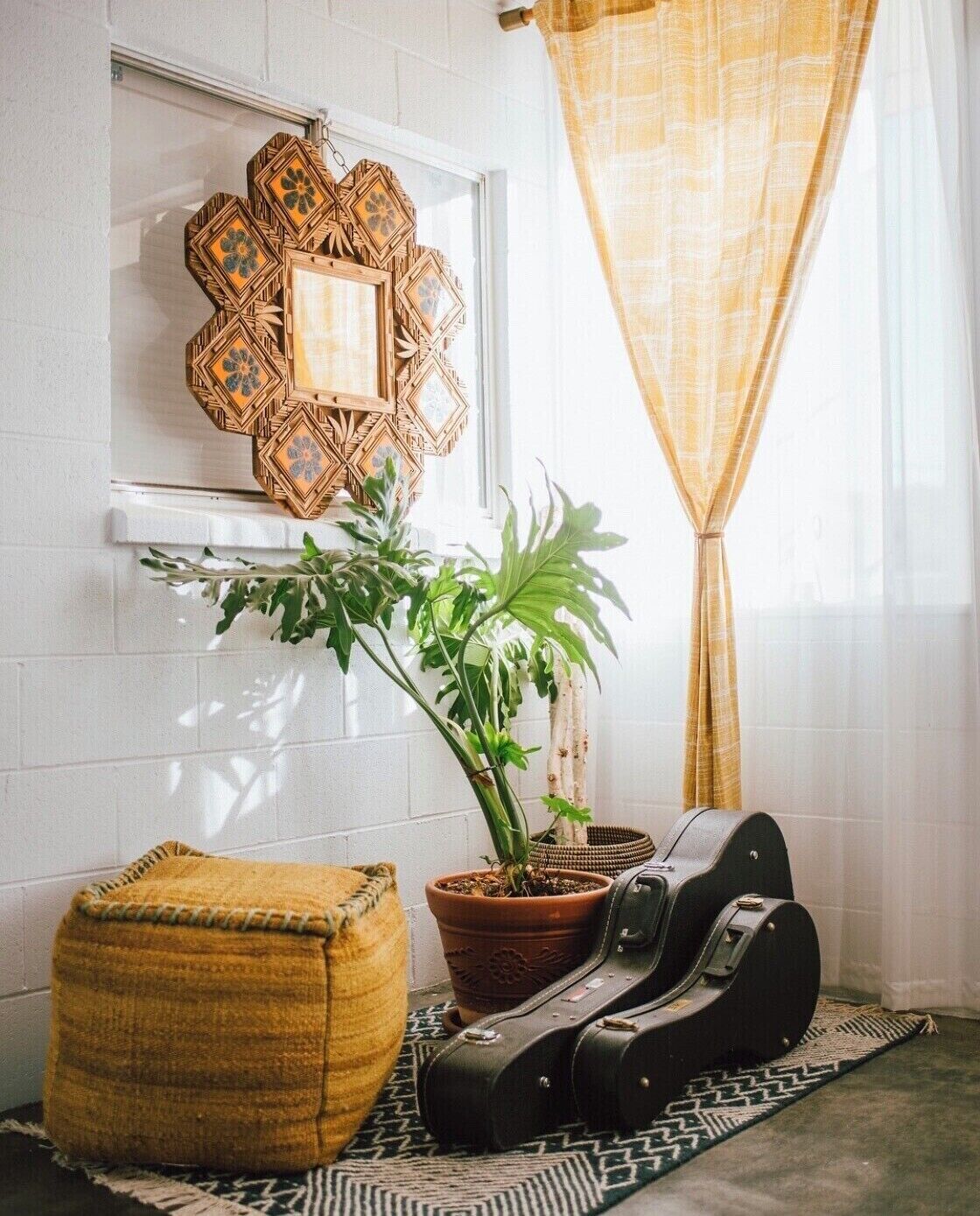 A room with a potted plant, a guitar, a woven pouf, and a decorative wall piece.