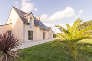 Ein modernes Haus mit Satteldach, Solaranlagen und einem gepflegten Rasen mit tropischen Pflanzen.