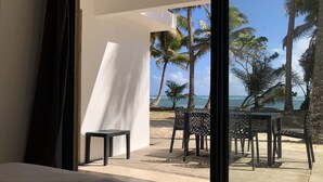 A room with a large bed, a view of a beach with palm trees, and a table and chairs outside. - Be On The Beach (Sainte-Anne)