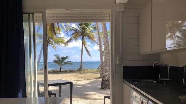 A kitchen with a washing machine, a table, and a view of the beach with palm trees and the ocean. - Be On The Beach (Sainte-Anne)