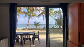 A room with a view of a beach, a dining table with chairs, and a large window. - Be On The Beach (Sainte-Anne)