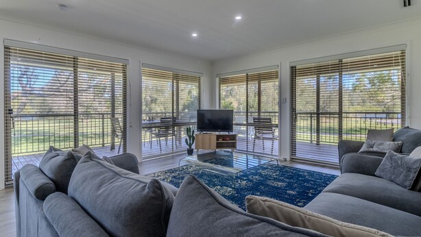 A spacious living room with a large grey sofa, a flat-screen TV on a wooden stand, and a dining area with a table and chairs.
