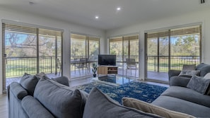 A spacious living room with a large grey sofa, a flat-screen TV on a wooden stand, and a dining area with a table and chairs.
