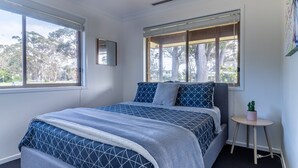 A bedroom with a bed, two windows, a small table with a plant, and a mirror on the wall.