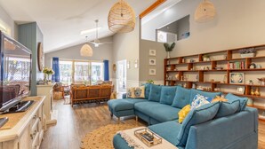 A spacious living room with a blue sectional sofa, wooden shelves with decorative items, and a large window providing natural light. - Fair Views over the Inlet (Sussex Inlet)