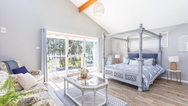 A bedroom with a four-poster bed, a sofa, a glass coffee table, and a view of a deck and trees. - Fair Views over the Inlet (Sussex Inlet)