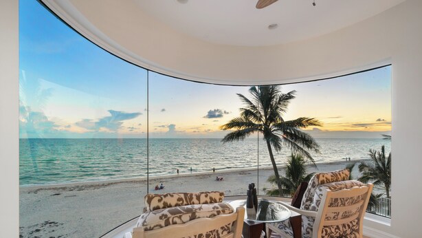 A room with a large curved window overlooking a beach, two patterned chairs, a small table with a bottle, and a ceiling fan. - Beachfront Vacation Home | Heated Pool | Panoramic Gulf Views (Bonita Springs)