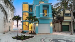 A modern two-story building with a light blue facade, a cylindrical glass tower, and a paved walkway with palm trees. - Beachfront Vacation Home | Heated Pool | Panoramic Gulf Views (Bonita Springs)