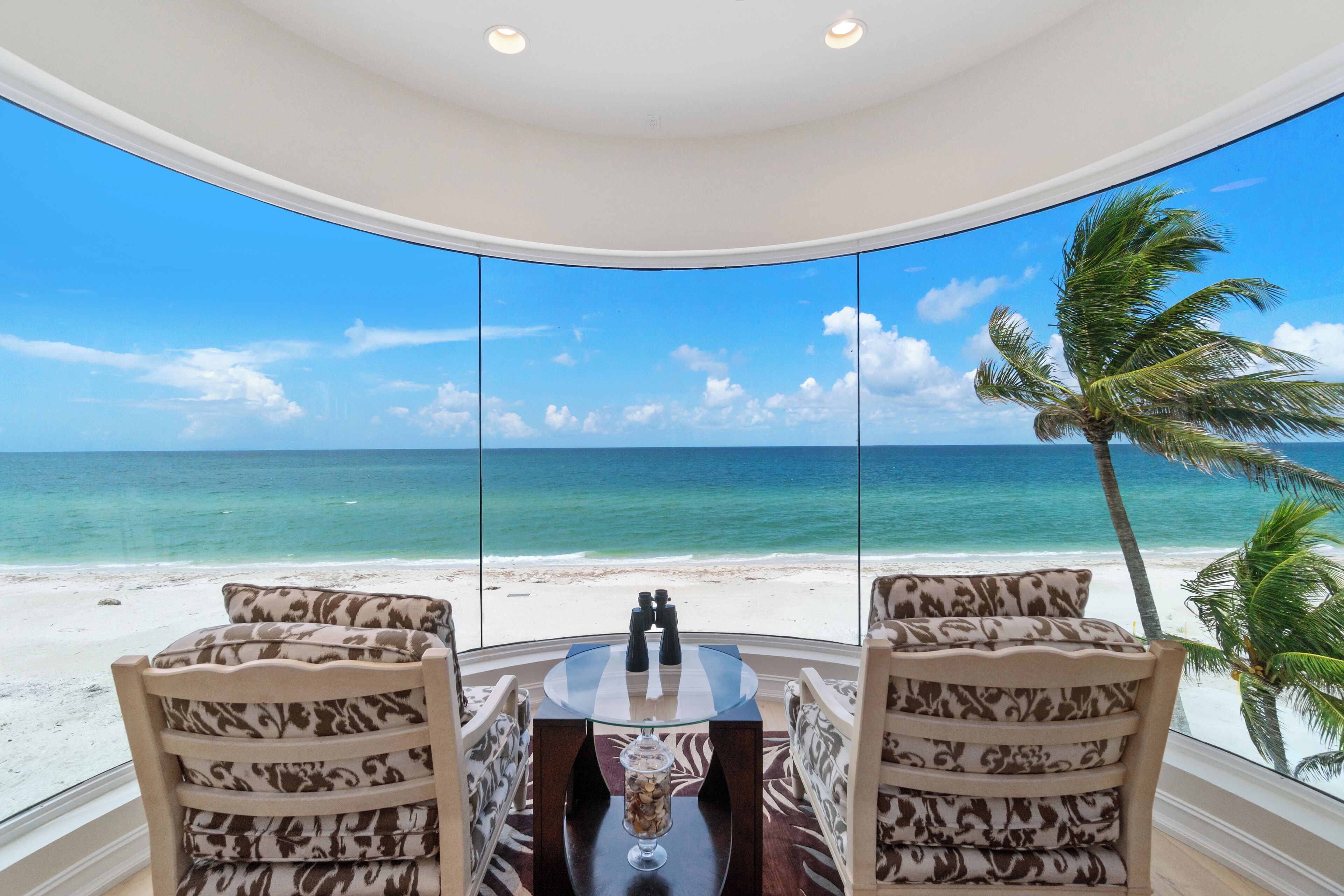 A room with a view of the beach and ocean, featuring a round table with chairs and a decorative centerpiece.