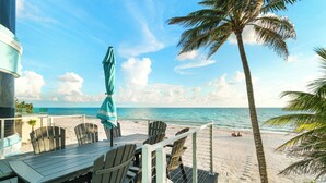 A wooden deck with a table and chairs, overlooking a beach with palm trees and the ocean. - Beachfront Vacation Home | Heated Pool | Panoramic Gulf Views (Bonita Springs)