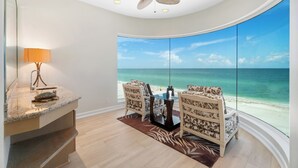 A room with a large window overlooking a beach, a ceiling fan, and a dining area with chairs and a table. - Beachfront Vacation Home | Heated Pool | Panoramic Gulf Views (Bonita Springs)