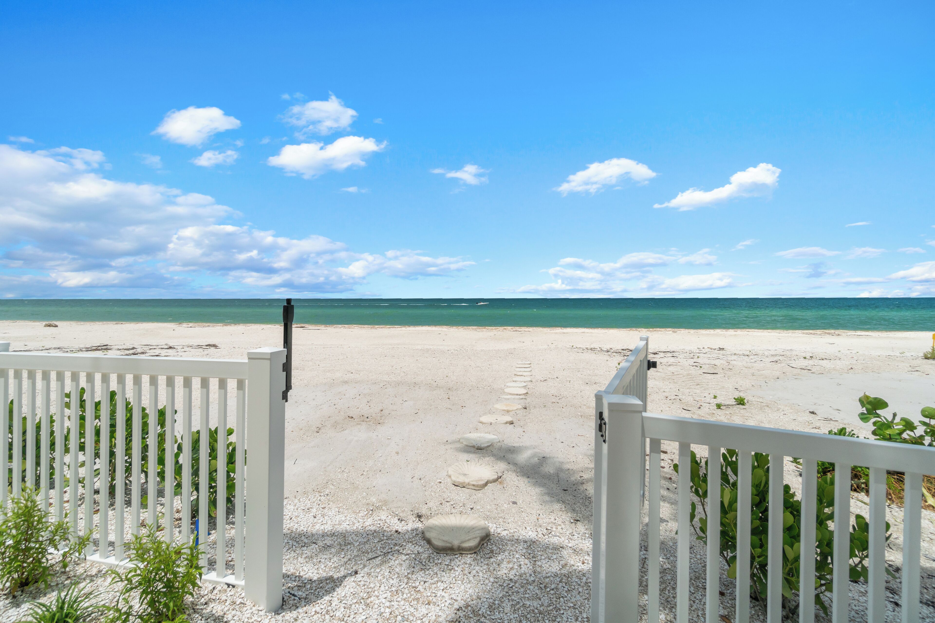 A beach view with a white fence leading to the water, clear skies, and calm sea.