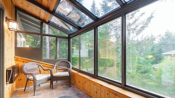 Ein Wintergarten mit großen Fenstern, Korbmöbeln und einer Holzbank.