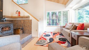 Ein gemütliches Wohnzimmer mit Kamin, einer Couch, einem gemusterten Teppich und großen Fenstern mit Blick auf den Wald.