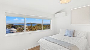 Un dormitorio con un ventanal que ofrece vistas de un paisaje costero, una cama con ropa blanca y un equipo de aire acondicionado instalado en la pared.