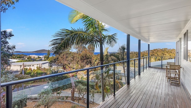 Una terraza de madera con barandilla de cristal que da a un paisaje costero con palmeras y vista al mar.