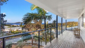 A wooden deck with a glass balustrade overlooking a coastal landscape with palm trees and a view of the sea. - Island View - 80 Lentara St - Large Family Home, Pool, WIFI and Sweeping Views of Fingal (Fingal Bay)