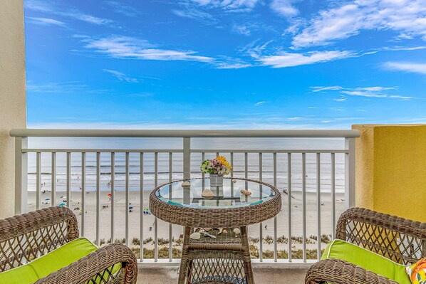 Ein Balkon mit Korbmöbeln, einem Glastisch mit Blumenvase und Blick auf den Strand.