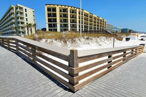 A beachfront hotel with a curved facade, multiple balconies, and a wooden boardwalk leading to the beach.