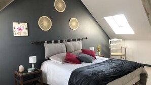 Un dormitorio con techo inclinado, cama con ropa de cama blanca y varios almohadones, mesita de noche con lámpara y un jarrón, y un tragaluz.