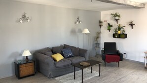 Un salón con un sofá gris, una mesita, un sillón rojo, una lámpara de pie y varias plantas en macetas sobre estantes.