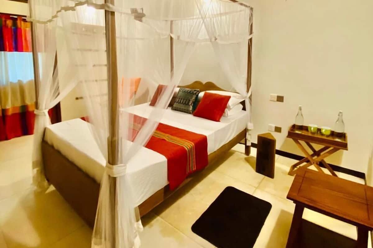 Ein Himmelsbett mit Baldachin, ausgestattet mit einer weißen Bettdecke und roten Akzenten, ein Nachttisch aus Holz sowie ein kleiner Tisch mit Flaschen.