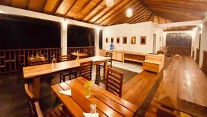 Un comedor interior con mesas y bancos de madera, un techo alto con vigas de madera y vistas al exterior de noche.