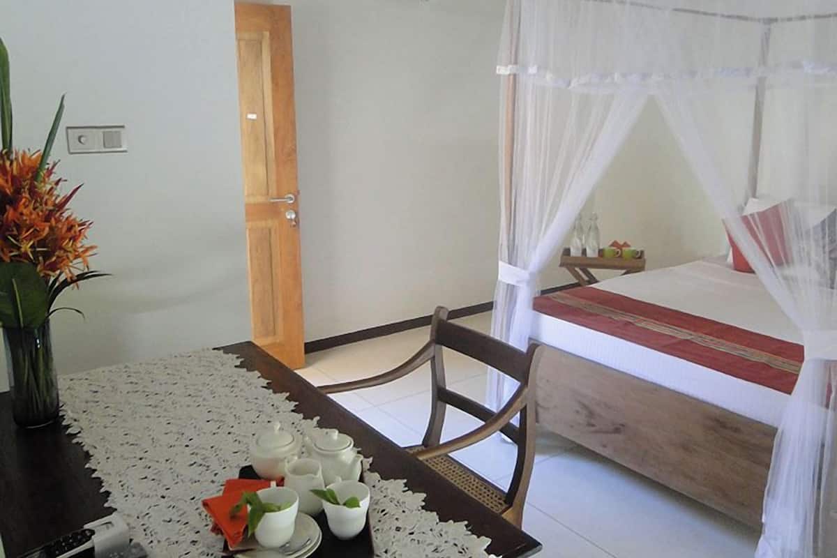 Ein Zimmer mit einem Bett unter einem Moskitonetz, einem Tisch mit einer Spitzenleinentischdecke und einer Vase mit Blumen.