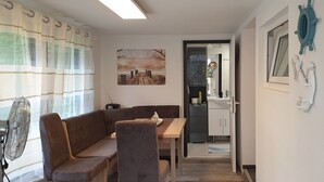 A cozy living room with a brown sofa, a wooden dining table, and a fan. A painting hangs on the wall, and a door leads to a kitchen area. - Great bungalow on the water (terrace, garden, beach, boat, bicycles, etc.) (Groß Kreutz (Havel))