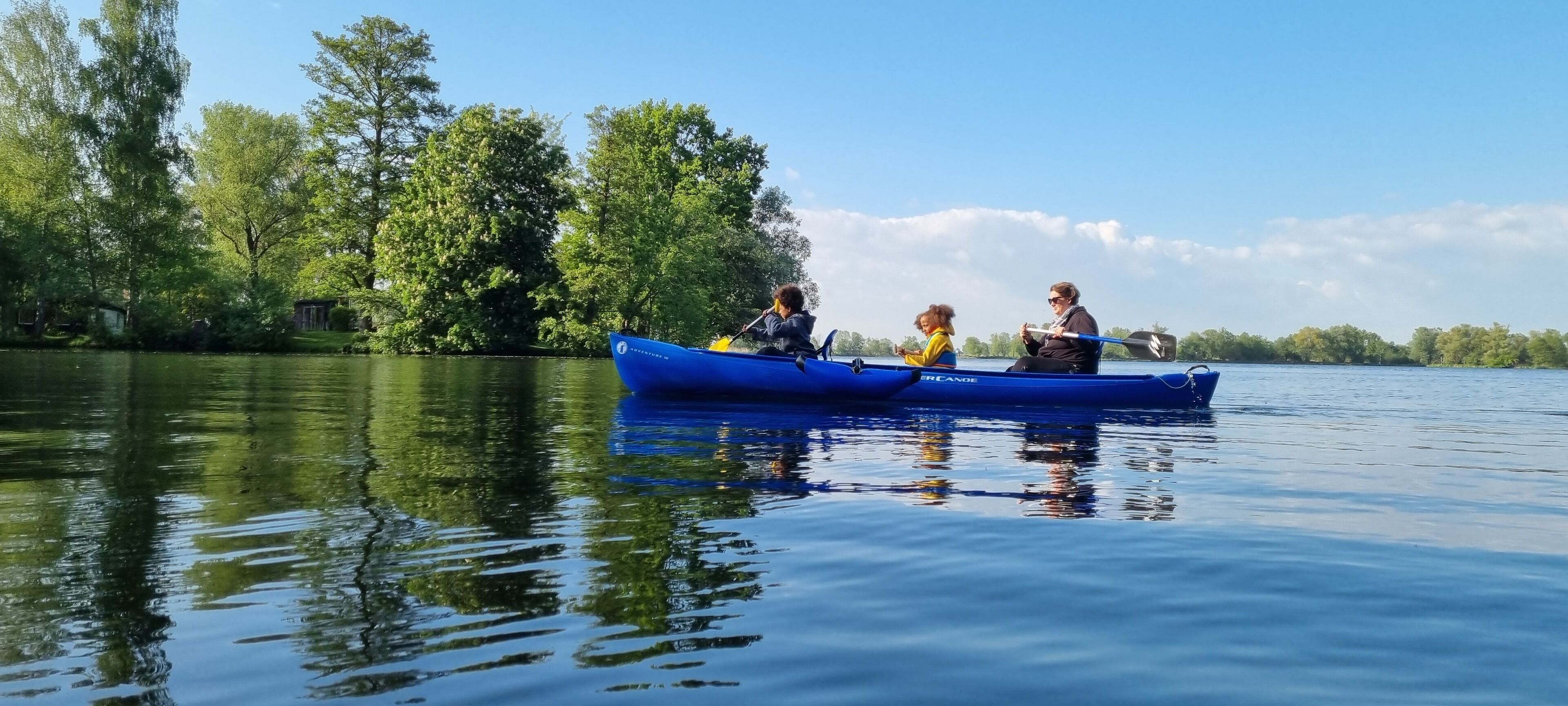 Un canoë bleu sur une eau calme, avec des personnes qui pagayent, entouré d’arbres et sous un ciel dégagé.